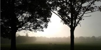 Image shows the early morning sun rising over Roundhay Park in Leeds with people walking in the distance