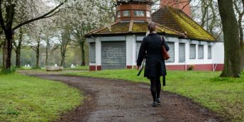 Woman walking alone on a curved path in a park toward a small, closed pavilion with shuttered windows