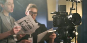 On a TV set, two workers discuss a script and storyboard behind a camera.