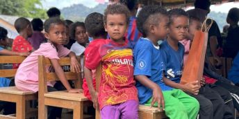 A group of Indigenous Jahai children dressed in colourful clothing sit together on chairs looking at the camera