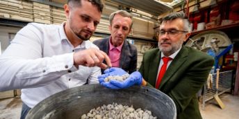 Three researchers looking at a container full of small pieces of recycled concrete. One researcher holds some in his hand. They are standing in an industrial-looking lab with exposed pipework and equipment behind them.