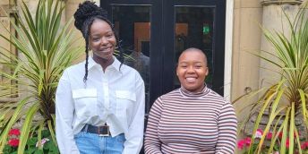 Two black women stand in front of green plants and open doorway smiling.