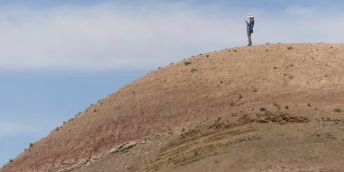 Person stands on steep brown hill against blue sky