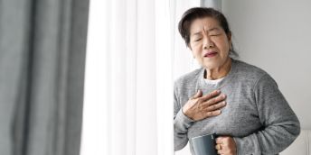 A person standing next to a window holding a mug in one hand and clutching their chest with the other hand