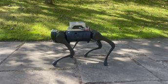 Four-legged, dog-type robot walking on concrete paving slab with grass area behind.