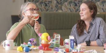 Two people sitting at a table covered with an array of dental-health related products. One of the people is holding a picture of a mouth in front of their mouth and is pretending to brush the teeth on the picture.