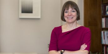 Professor Shearer West standing in an office, smiling and wearing a purple top, her arms folded