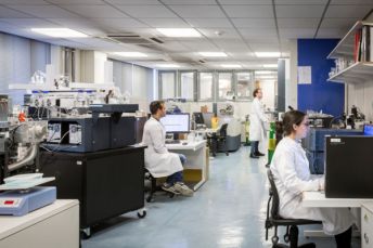 3 researchers work inside a large mass spectrometry lab. The researchers are wearing white lab coats and working in different parts of the lab, including working on computers and checking ongoing experiments.