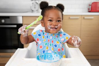 A baby girl sitting in a highchair smiling and holding a spoon