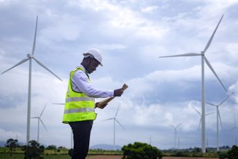 A person in a high vis vest and hard hat stands in front of wind turbines looking at a clipboard