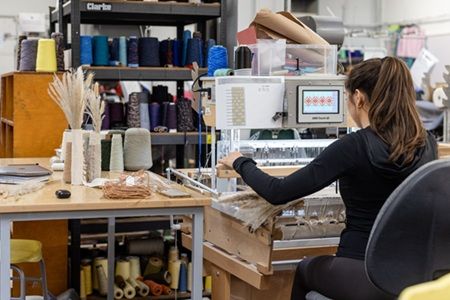 A person sat in a studio space operating some textile weaving machinery and computer software.