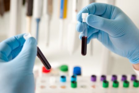 Two gloved hands holding test tubes of blood in a laboratory.