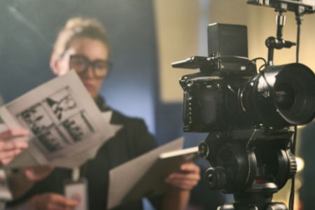 A member of a camera crew stands behind the camera, looking at storyboards.