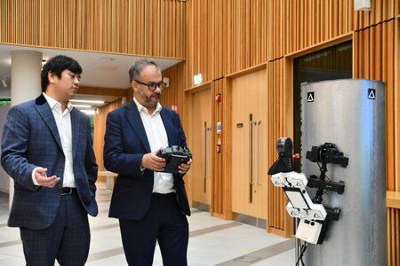 The Minister for Tech and Digital Economy has a remote controller in his hand controlling a robot which is on a tube in front of him. A member of the Acuity Robotics team is stood beside the Minister telling him how to control the robot.
