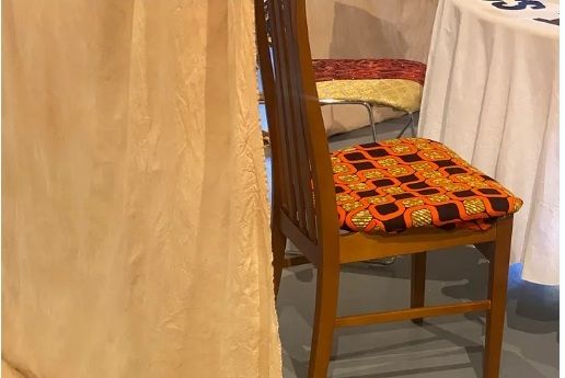 A table with tablecloth and two dining room chairs upholstered in patterned fabric, with a large curtain partially obscuring the view.