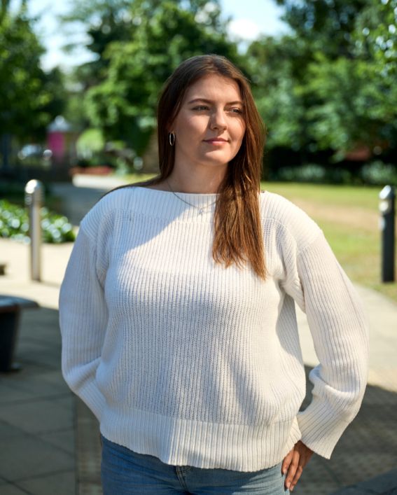 Student Katie Hartmann stood on campus by a green space with trees and bushes.
