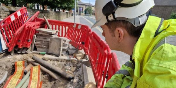 Alex Sunderland wears a yellow hi-vis coat and a hard hat as he looks at down at road works surrounded by red barriers in Leeds City Centre.