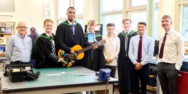 A group of graduates holding their guitar device which is fixed to the guitar with Alex Sobel MP, two professors and REMAP client.