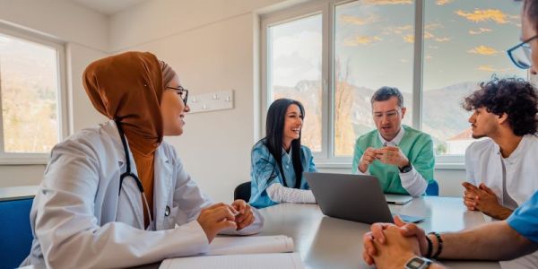 A group of medical professionals sit around a table talking and looking at a laptop