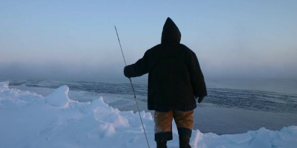 A hooded figure is seen from the rear as they stare at the Arctic ice and the sea.
