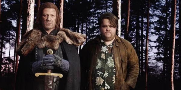 Danny Kirrane stands in a forest beside Sean Bean who is holding a sword, dressed as the character Ned Stark from Game of Thrones