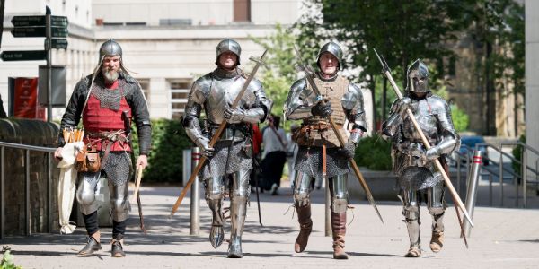 A group of four reenactors dressed in armour walk towards the camera in a row, on Beech Grove Plaza at the University of Leeds.