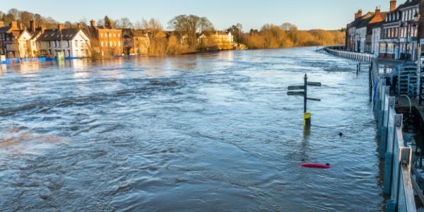Flooded riverside town with fast-moving brown water covering a riverside path and partially submerging signposts, with rows of brick and white houses along both banks under clear daylight.