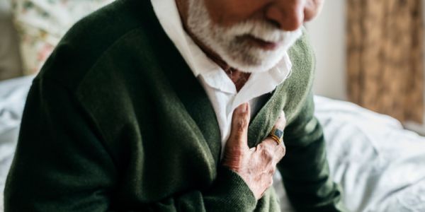 An elderly person with a white beard and wearing a green cardigan clutching their heart