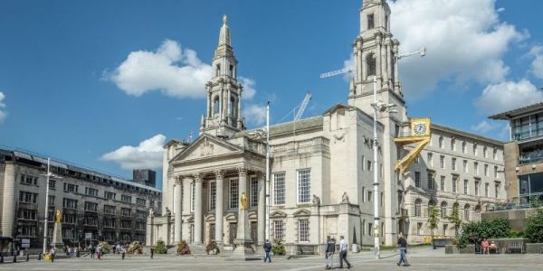 A grand stone civic building with tall twin clock towers, large arched windows, and a columned entrance stands under a bright blue sky with scattered clouds. The open square in front of the building has a few people walking across wide paved area.