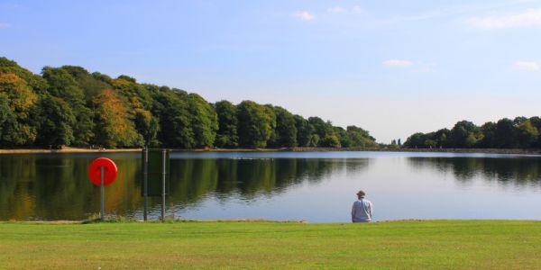 A figure stands by the lake in Roundhay Park in Leeds. The lake is reflecting the bright blue sky.