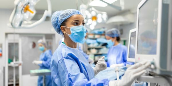 Healthcare professional in surgical gown, mask and gloves operating a medical monitor in a hospital operating theatre, with other clinical staff and equipment visible in the background.