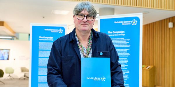 Simon Armitage holds a blue folder with the Yorkshire Cancer Research logo and looks at the camera.