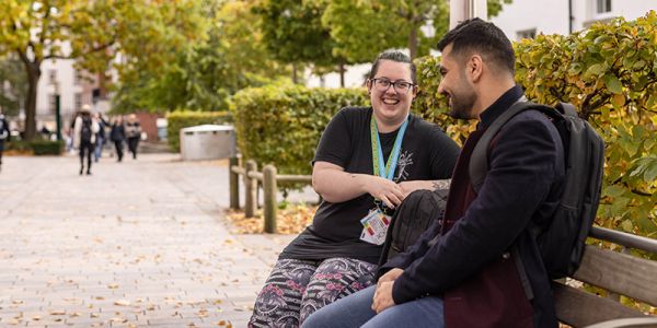 Two people sat on a bench on campus, smiling. There are leafy trees beyond.