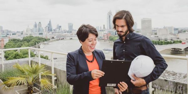Two individuals stood on a balcony with a cityscape behind them, looking at a computer tablet.