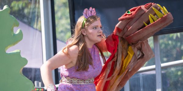 An actor wearing a purple dress and flower headband on stage, holding a dragon puppet with her left hand.