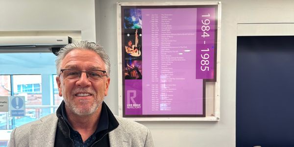 Stuart Galbraith stood in front of a gig listings poster for 1983 to 1984 in the University Refectory