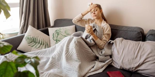 A person sitting on the sofa with a cup of tea and a blanket, looking exhausted