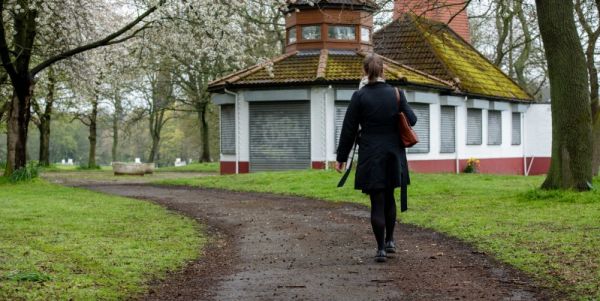 Woman walking alone on a curved path in a park toward a small, closed pavilion with shuttered windows