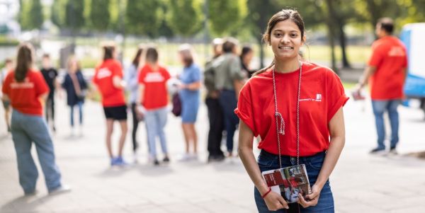 A student ambassador smiling at the camera, with other student ambassadors and visitors in the background, at an open day.