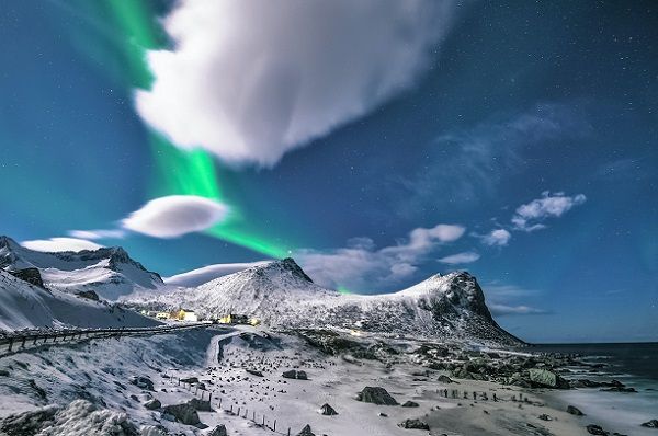 A snow capped mountain with clouds and the northern lights against a blue sky