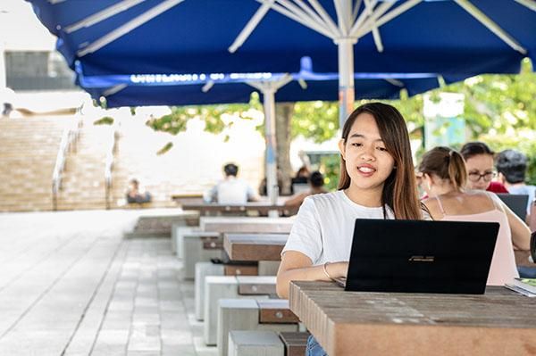 A student sitting outside under an umbrella using a laptop.