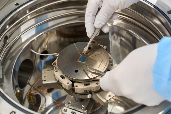 Two hands wearing white nitrile gloves use metal tweezers to place a small square silicon piece on a round metal disk at the centre of a cylindrical metal chamber.  