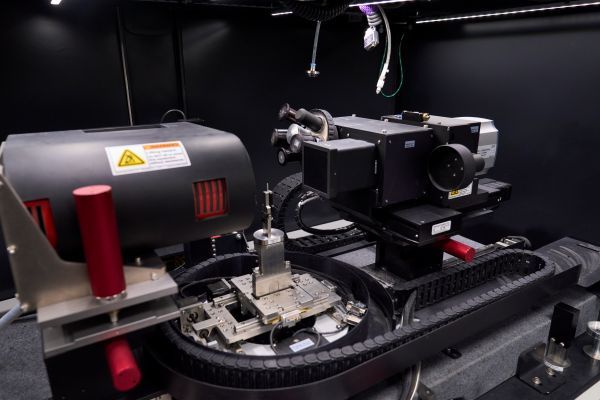 The inside of the XCT is shown with a thin, cylindrical sample mounted in a clamp between an X-ray source and an X-ray detector.  The sample is on a rotating stage.