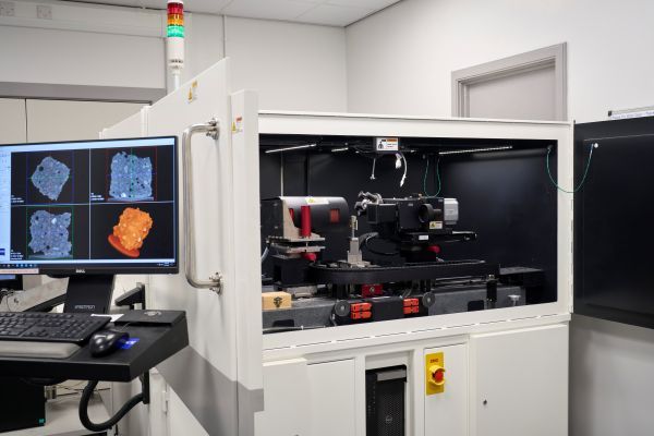 The open XCT, a large white metal chamber.  A porous, concrete sample is mounted between an X-ray source and an X-ray detector.  A computer monitor outside of the machine displays 2D and 3D imaging data for the sample.