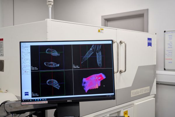 The closed XCT, a large white metal chamber with Zeiss branding visible on its doors. A computer monitor outside of the machine displays 2D and 3D imaging data of a portion of a rabbit skull. 