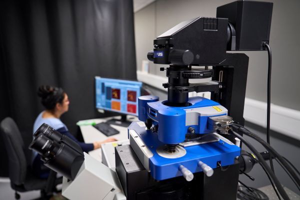 A close-up of the blue and black microscope, with a person seated at a computer reviewing images on a monitor in the background.
