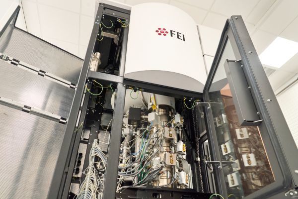The microscope, a room-height metal and glass structure stands with its door open, revealing wiring and mechanics. The structure is branded with the FEI logo.