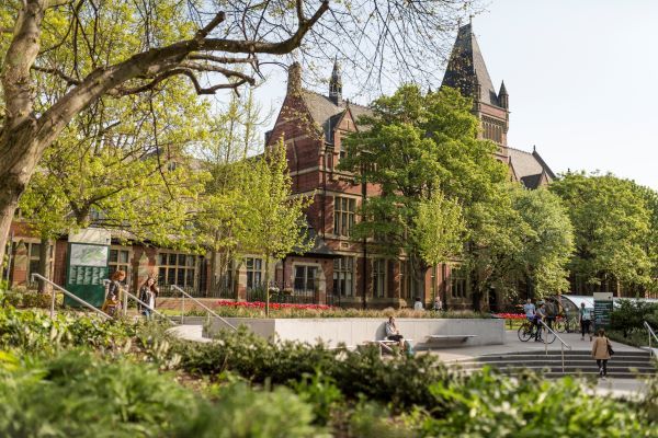 The Great Hall, a big redbrick university building, is photographed through blooming trees on a sunny day on campus.