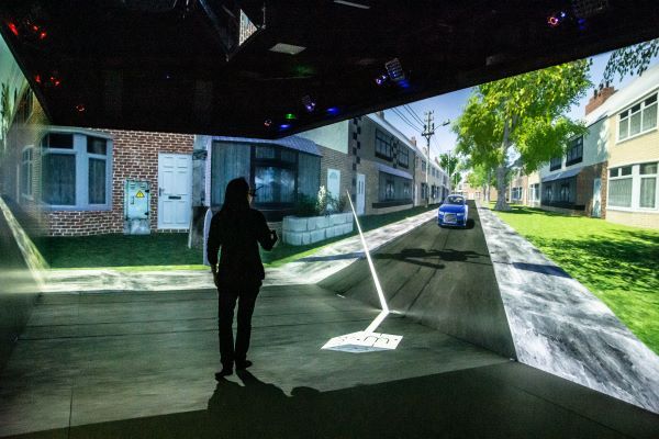 A person is silhouetted against a bright virtual reality display, wearing VR glasses and interacting with in a simulation of a suburban street. The projection on the walls depicts houses, a car driving down the street, and greenery along the pavement.