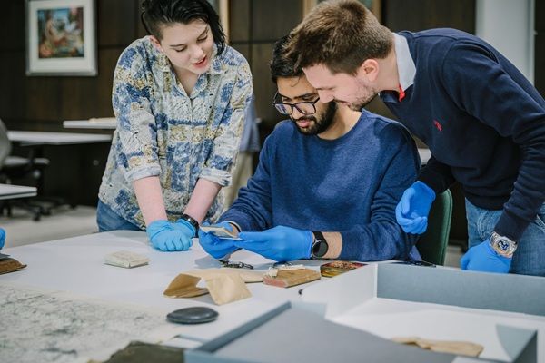 A group of three researchers carefully examine historical items from an archival collection, all wearing blue gloves to preserve the fragile materials.